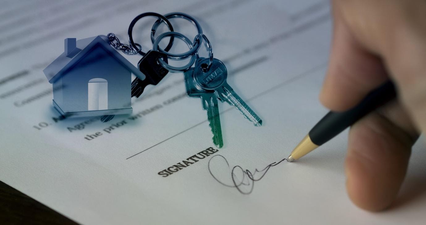 Image of a document which is being signed by a person only the hand and pen is visible There is a set of keys with a house shaped key right resting on the paper which is for an investment property