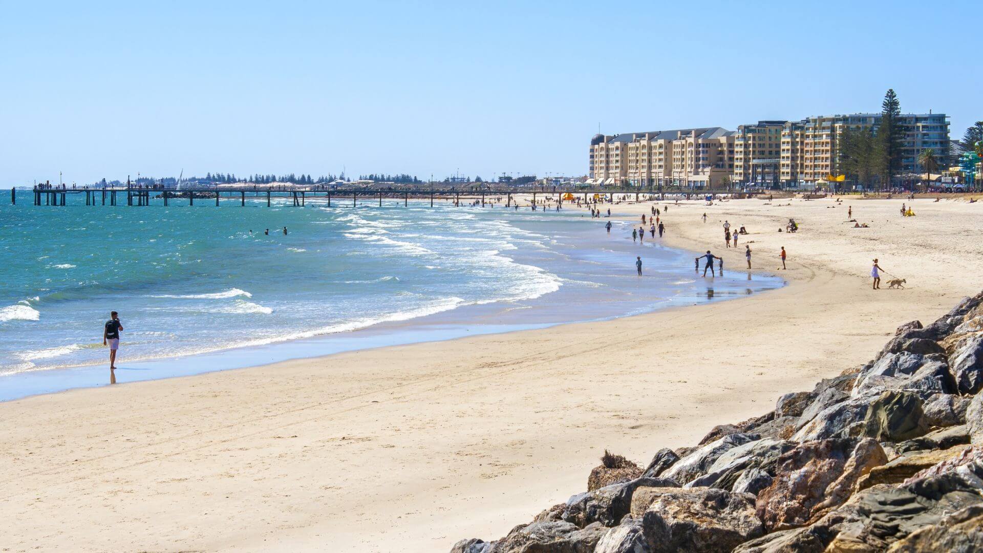 A bustling beach scene at Glenelg, South Australia, with golden sands meeting gentle waves. People enjoy various activities, from walking to playing, under a bright blue sky. In the background, the Glenelg Pier extends into the sea, while a row of coastal apartment buildings lines the shore, reflecting the popular and vibrant seaside suburb's lifestyle.