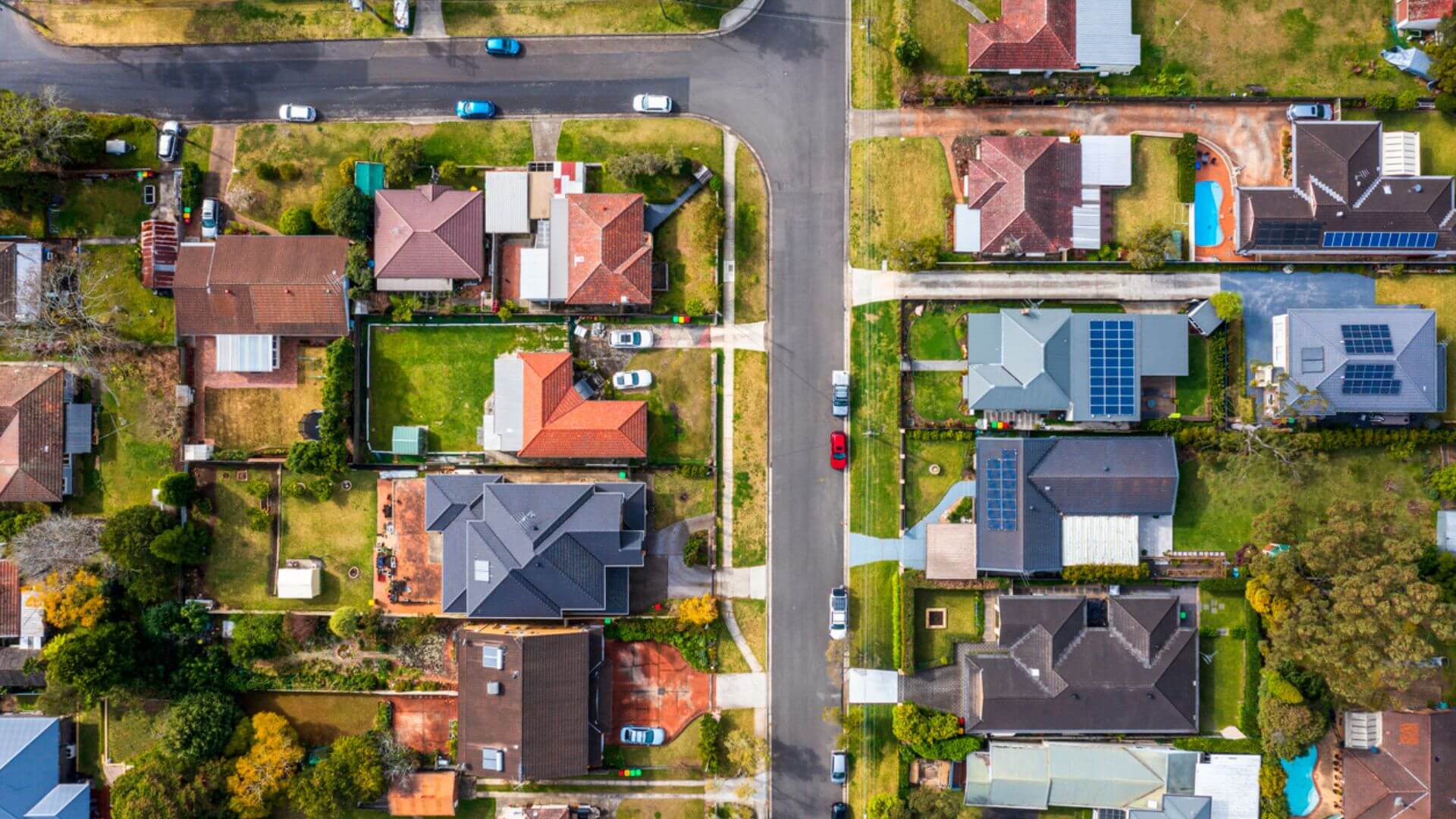 This is an aerial view of a suburban neighborhood showing a variety of detached single-family homes with different roof designs and various shades of roofing materials. The image captures a section of a residential area with a street running vertically through the middle. There are visible front and back yards, some with lush green lawns, gardens, and a few trees. Several houses have driveways with parked cars, and one house on the right features a swimming pool. The presence of solar panels on a few rooftops suggests a community with energy-conscious residents. The overall pattern of the neighborhood layout is clear, with each property distinctly separated by fences or vegetation.