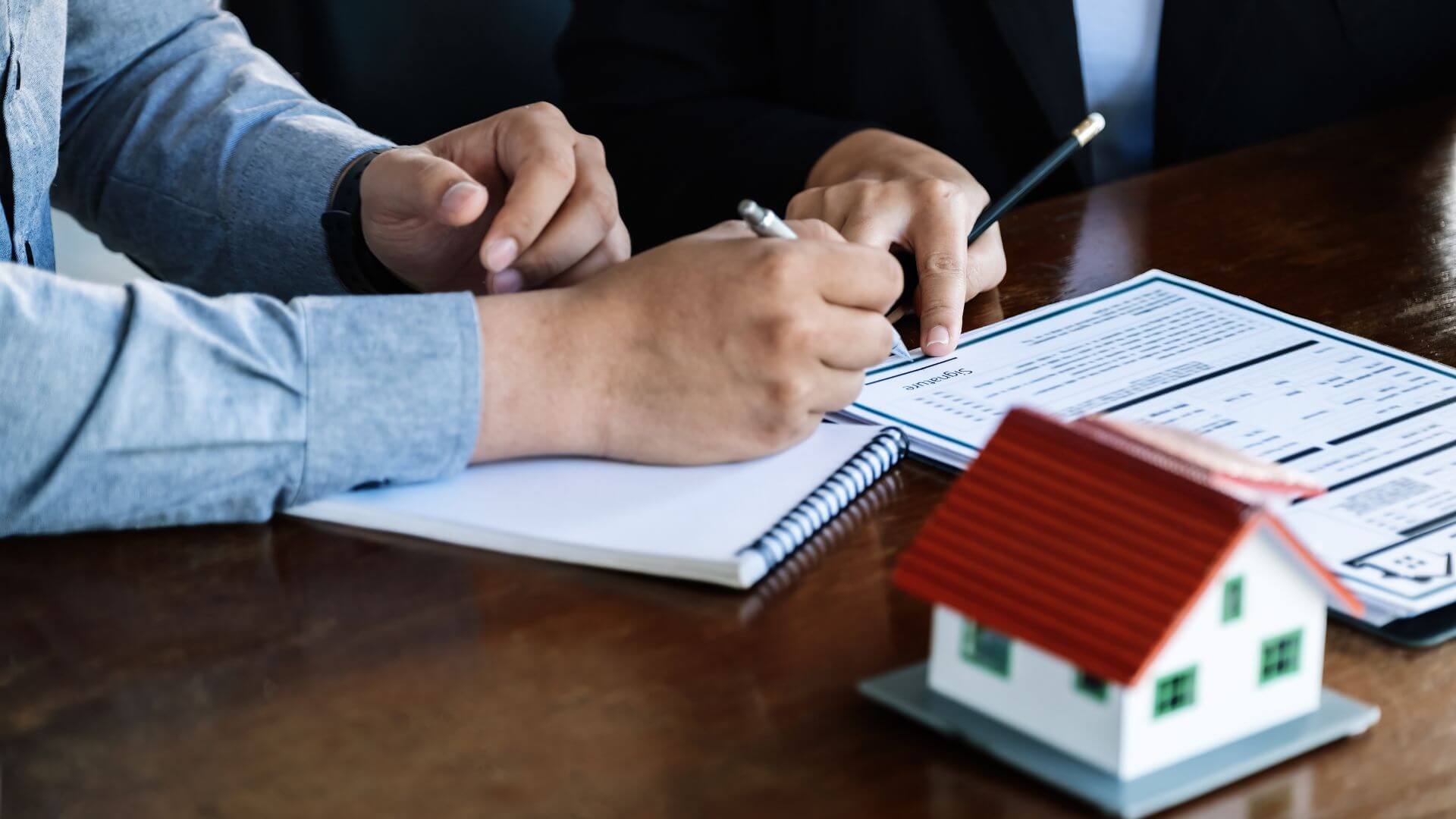 Two individuals in a professional setting are reviewing and signing documents with a focus on refinancing and researching as property investors. In the foreground, there's a miniature model of a white house with a red roof, symbolising property ownership or investment. The scene suggests a meeting between a client and a real estate professional or a financial advisor, discussing property-related finances or contracts.