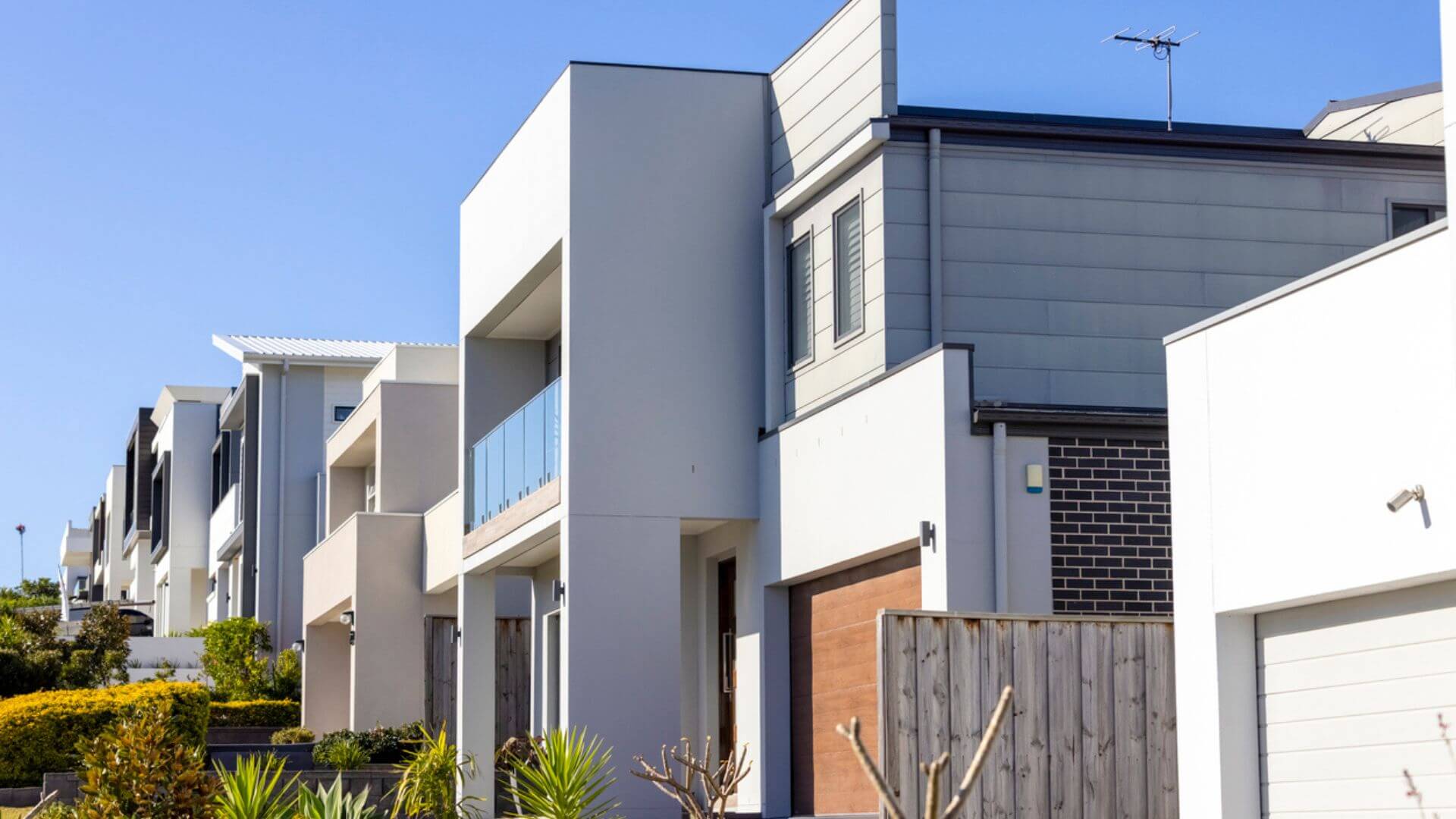 A row of modern, two-story townhouses with a mix of white and grey facades under a clear blue sky. Each home features a balcony, large windows, and a garage, complemented by neatly landscaped front gardens with lush shrubs and a manicured lawn. A television antenna is visible on the roof of one townhouse.