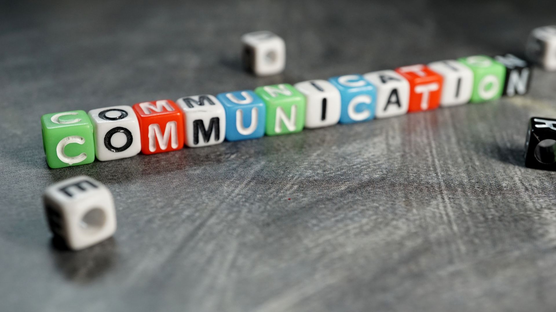 Colourful lettered dice arranged on a grey fabric surface to spell out the word 'COMMUNICATION' in a diagonal line. The dice feature a variety of vibrant colors like green, red, blue, and white, with each cube displaying a single letter. The image conveys the concept of building effective communication, essential in various contexts such as business and personal relationships.