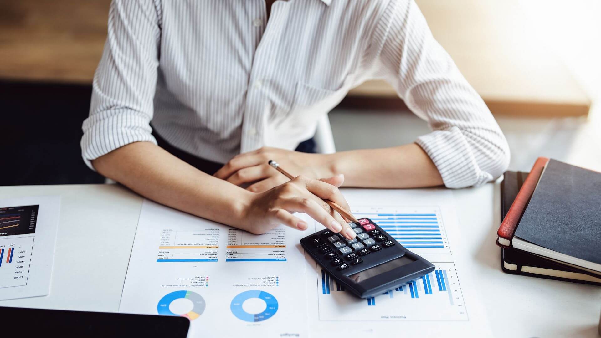 A person sits at a desk analyzing financial charts and data with a calculator in hand, indicative of meticulous financial planning or budgeting. The setting suggests a thorough review of investment performance or preparation for a financial strategy, with notebooks and a laptop nearby, ready for detailed work.