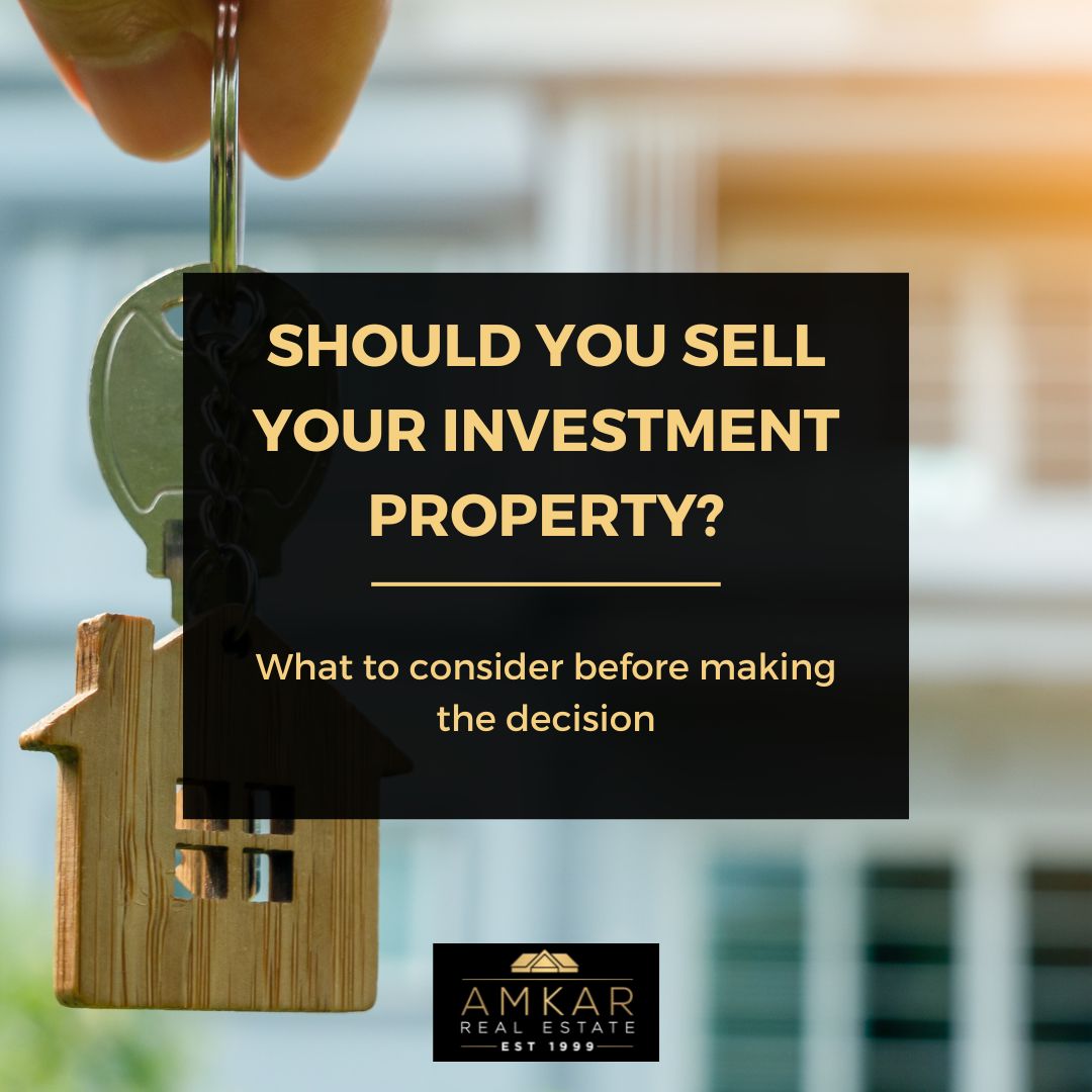 The image features a square graphic with a clear and modern design. The top half shows a hand holding a key with a house-shaped keychain, symbolizing property ownership. The background is an out-of-focus image of a residential home. The bottom half has a black background with the text "SHOULD YOU SELL YOUR INVESTMENT PROPERTY?" in bold white and gold letters, followed by "What to consider before making the decision" in smaller font. The AMKAR Real Estate logo is placed at the bottom, signifying the brand. The graphic likely serves as a visual prompt for a blog post or article related to making informed decisions about selling investment properties.