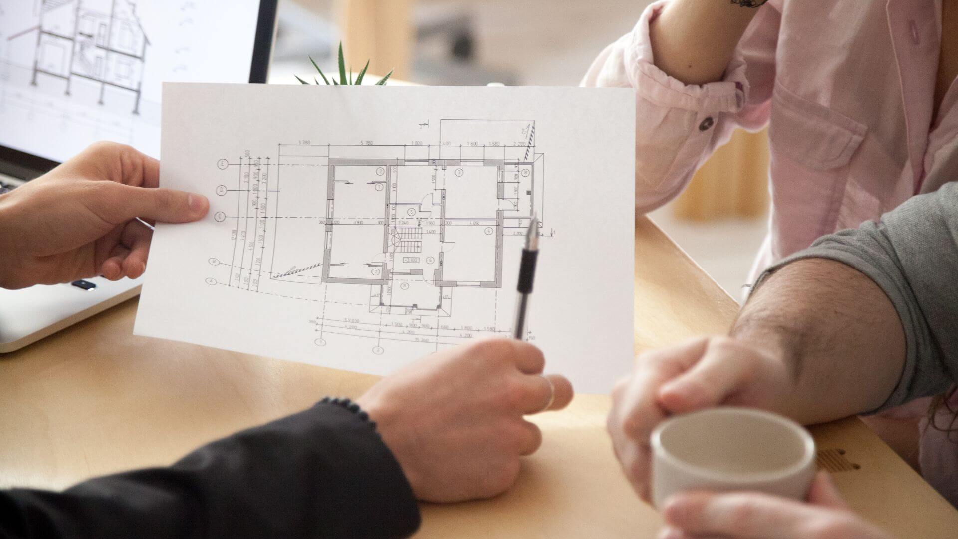 A close-up photo showing two individuals reviewing a detailed architectural floor plan. One person holds the blueprint steady on a wooden table, while the other, holding a pen, appears to be discussing specific aspects of the design. The blueprint is rich with technical measurements and layout details, suggesting a planning or consultation phase for property renovation or development.