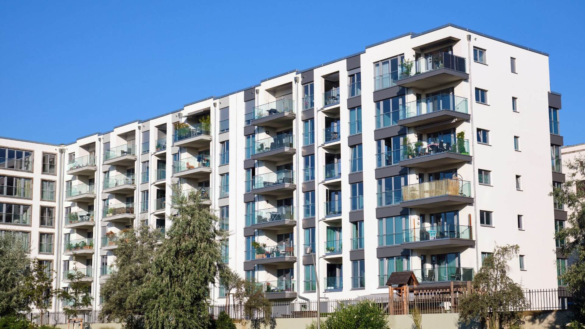 Modern multi-storey apartment complex featuring clean architectural lines, ample balconies with glass balustrades, and a variety of green plants enhancing the urban living space. The building is set against a clear blue sky, reflecting a contemporary urban lifestyle.