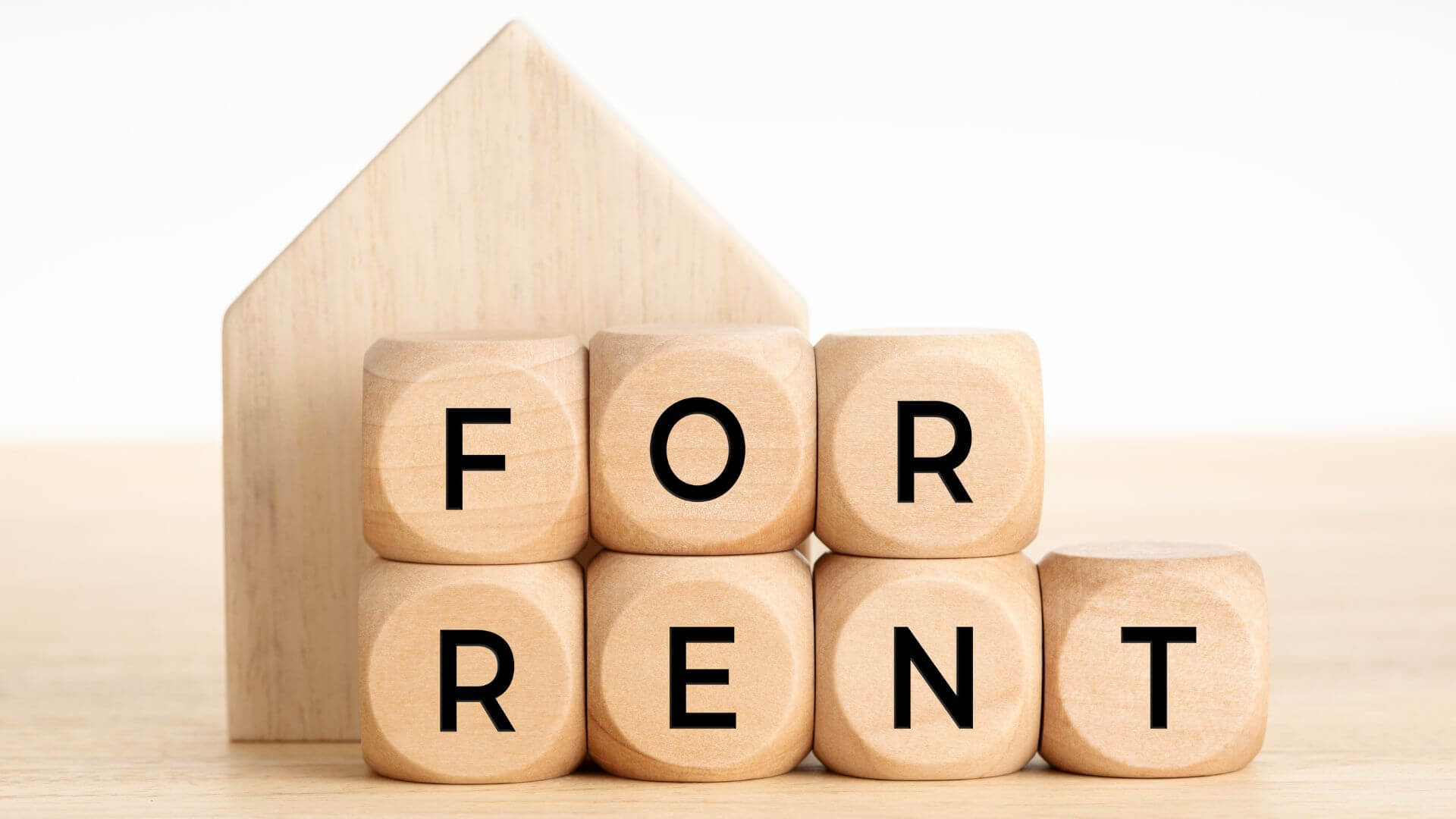 Wooden blocks spelling out 'FOR RENT' in front of a minimalist wooden house silhouette, symbolizing the considerations for setting rental prices in real estate. Key topics include market analysis, property features, local amenities, and finding the balance for competitive pricing.