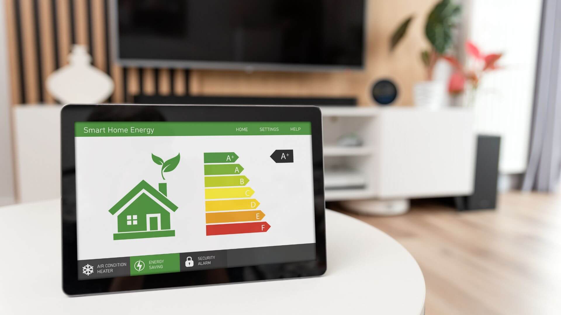A smart home energy monitor displayed on a tablet, showing an energy efficiency rating chart with a house icon and various energy classes from A+ to F, placed on a living room table with a TV and indoor plants in the background, emphasising a modern, energy-conscious home interior.