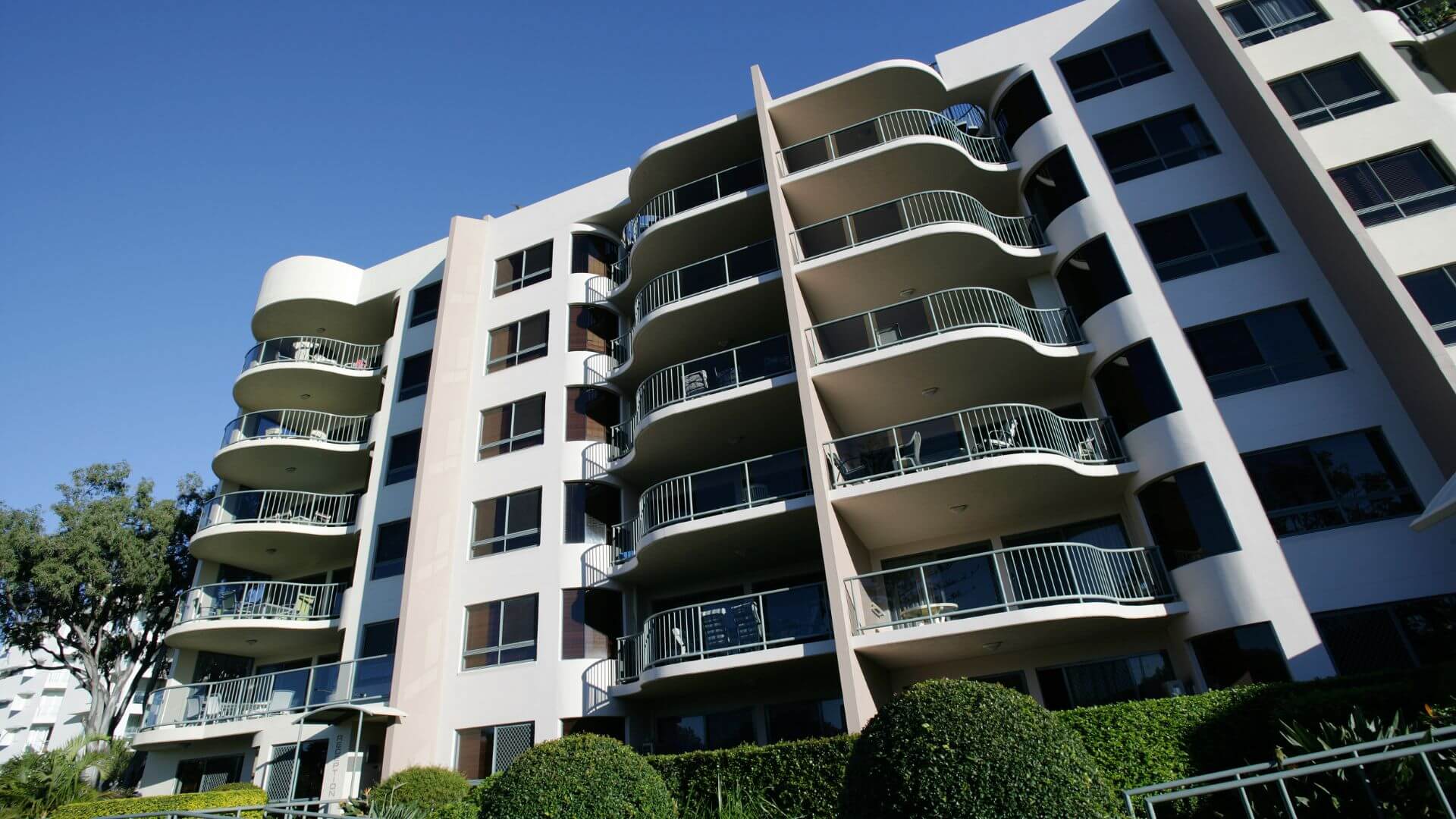 A modern apartment building bathed in sunlight, showcasing multiple balconies on each floor, with lush greenery at the base reflecting a comfortable, urban living space.