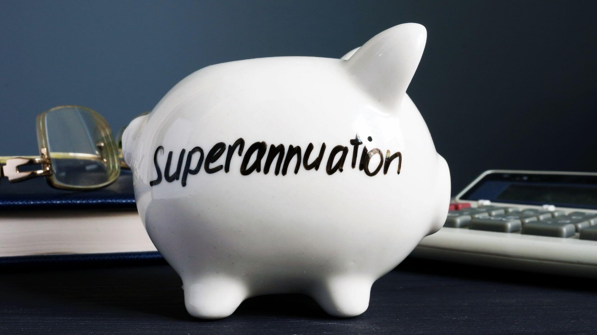 A piggy bank labeled "superannuation" sits in the foreground on a dark blue surface, symbolizing savings for retirement. Behind it, a pair of eyeglasses and a calculator atop a leather-bound book suggest financial planning and calculation, emphasizing the importance of preparing for future financial security.