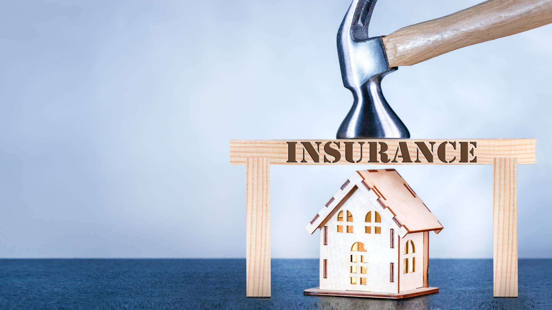 A wooden model of a house sits beneath a wooden block structure spelling out 'INSURANCE,' with a hammer positioned above as if to emphasize protection, set against a calm blue sea and sky background, illustrating the concept of landlord's insurance as a safeguard for property investment.