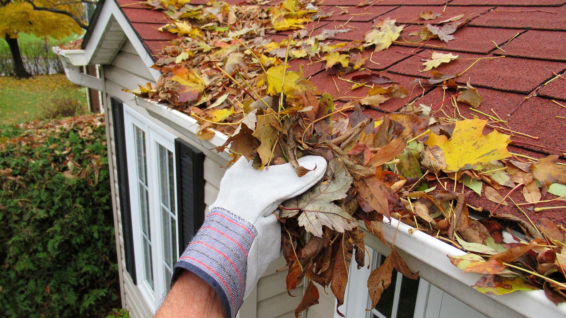 A person wearing gloves cleans leaves and debris from a house gutter as part of preparing the property for winter. This maintenance task helps ensure proper drainage and prevents damage during the colder months.