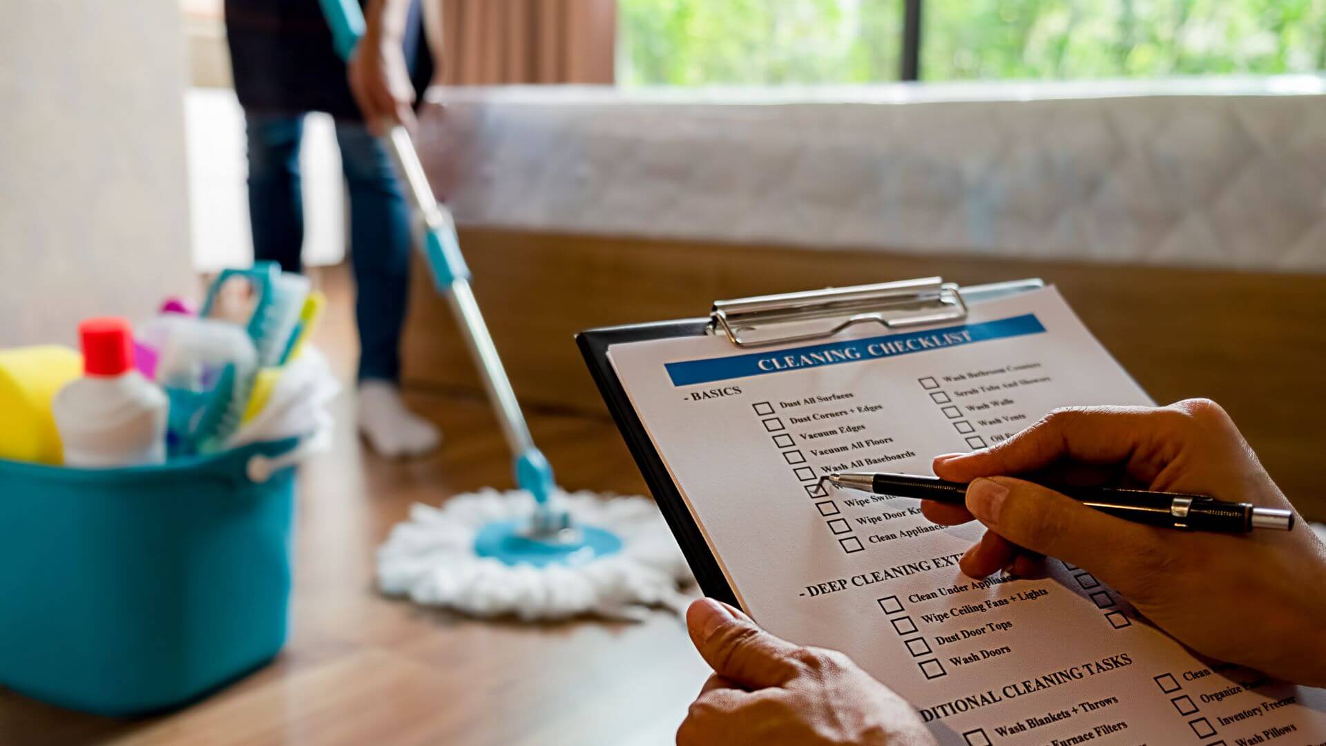 A person is holding a checklist titled "Cleaning Checklist," reviewing the tasks while another individual cleans the floor with a mop in the background. Cleaning supplies are visible in a bucket nearby, creating a clear focus on the importance of thorough end-of-lease cleaning to ensure all tasks are completed and the property is left in top condition.