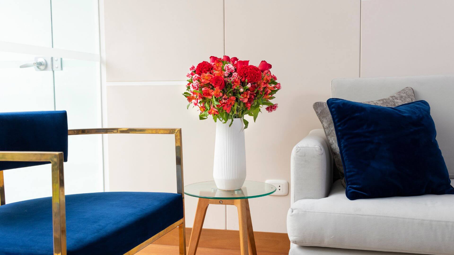 A cozy living room corner featuring a stylish navy blue chair with gold accents, a white vase filled with vibrant red and pink flowers on a glass table, and a light grey sofa adorned with blue and grey cushions.