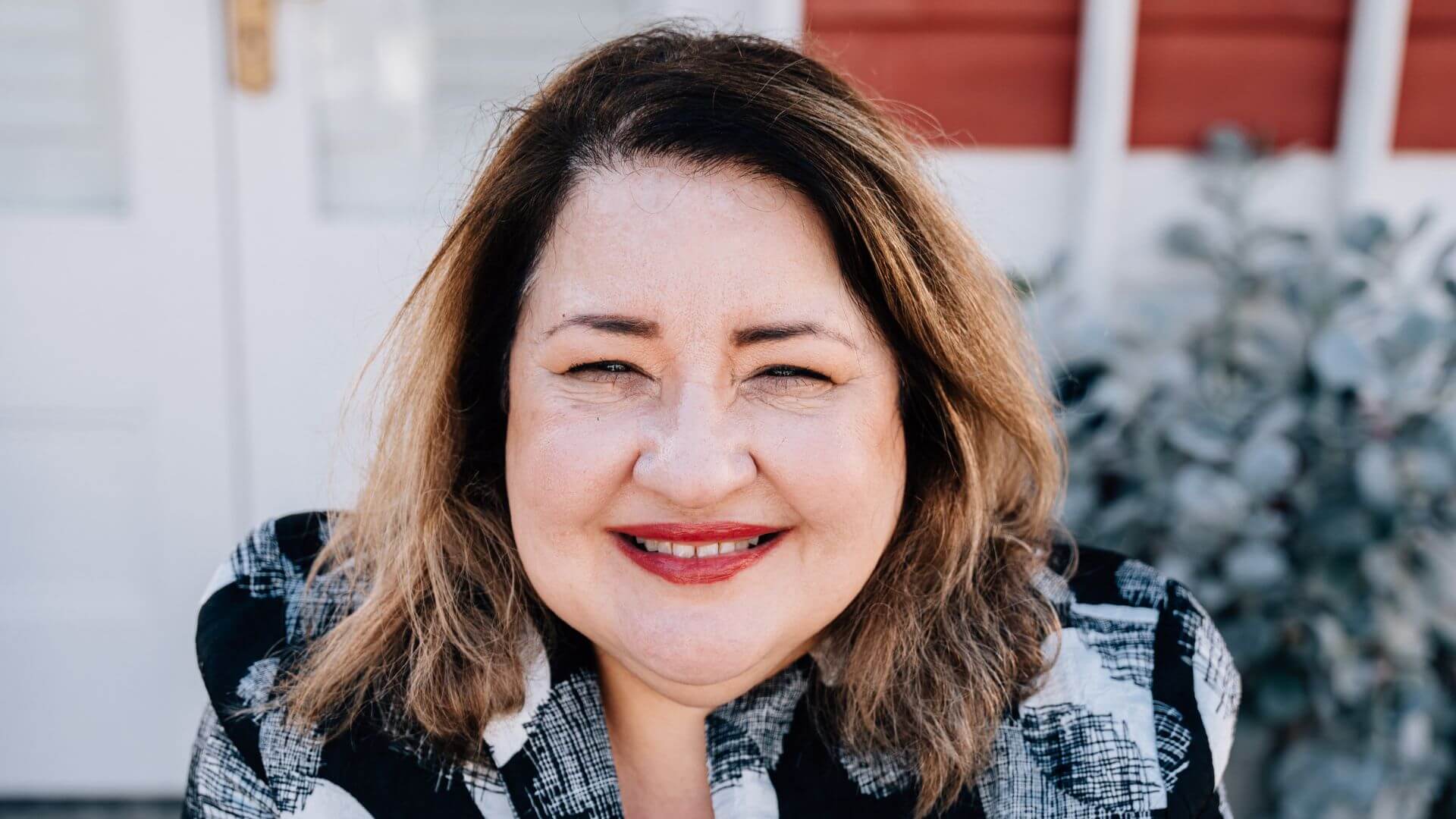 A portrait of Amal, a distinguished property manager and managing director, smiling warmly. Amal, known for her expertise providing support for people who are investing in property, is pictured outdoors with a backdrop of a white wooden fence and a leafy plant, illustrating her approachable and professional demeanor in the real estate industry.