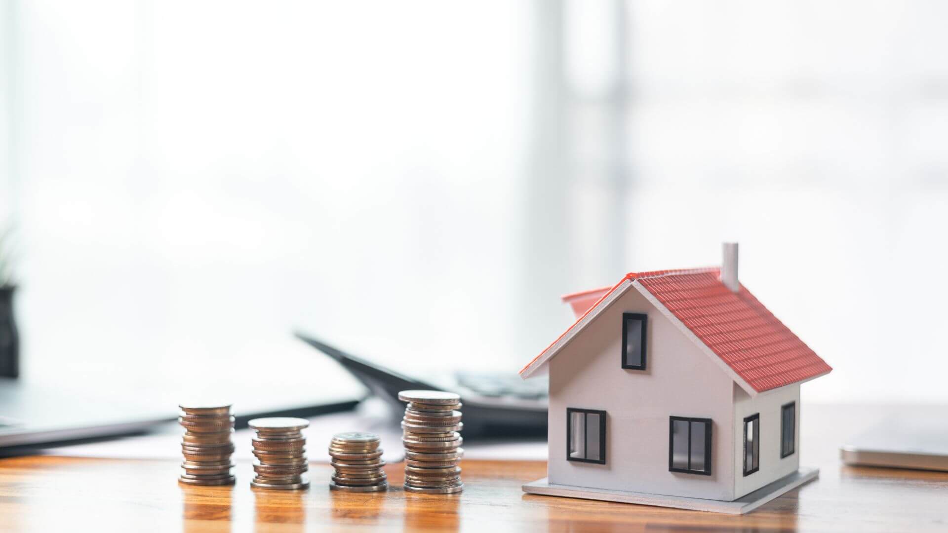 A small model house with a red roof sits on a desk next to stacks of coins, symbolising rental yield and property investment returns.