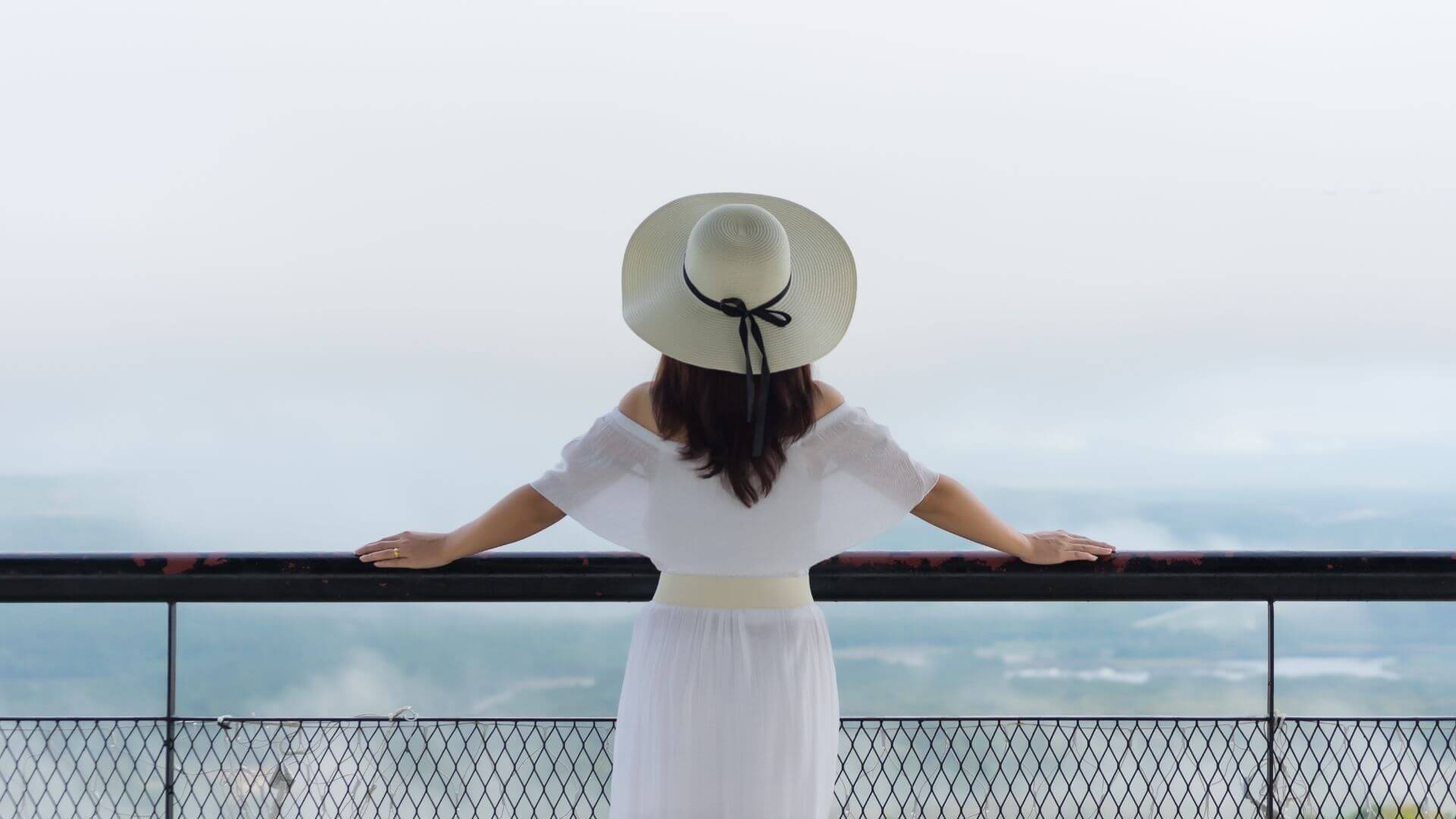 Person enjoying balcony living, standing at a railing with arms outstretched, wearing a white dress and wide-brim hat, overlooking a scenic view